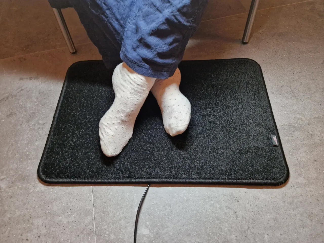 Zwarte verwarmde voetenmat Excellent op een tegelvloer: voetenwarmer voor thuis of onder het bureau